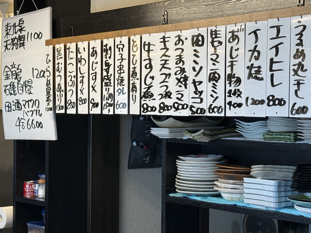 カウンター奥に下がった手書きの品書きの札。いわし酢〆、牛すじ煮、キス天ぷら、エイヒレなどが並ぶ