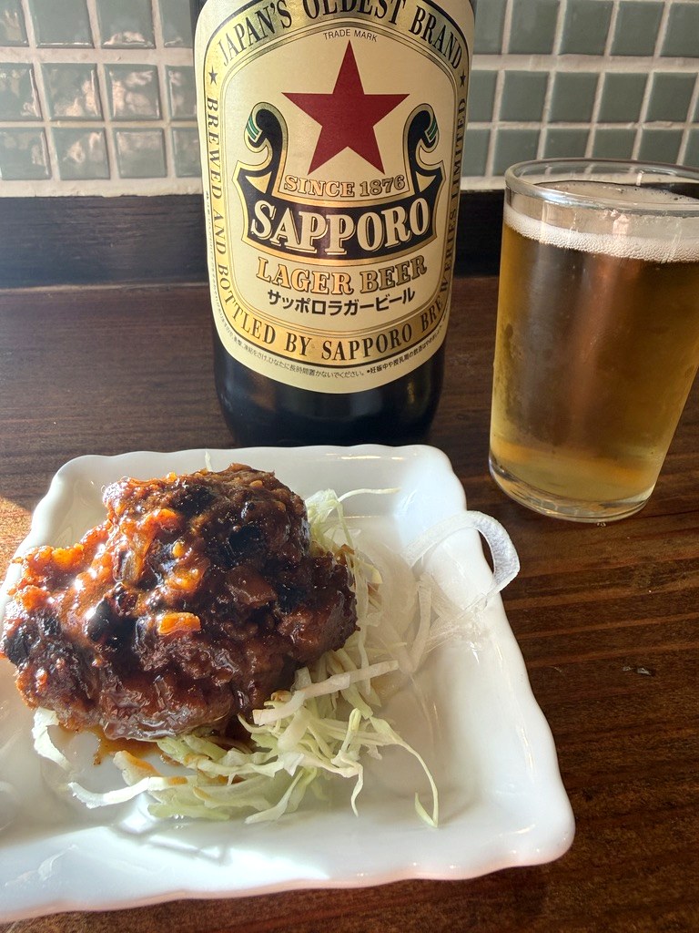 明るいうちのカウンター。サッポロラガービールの瓶とグラス、白い角皿の一品。奥に店の緑のタイル
